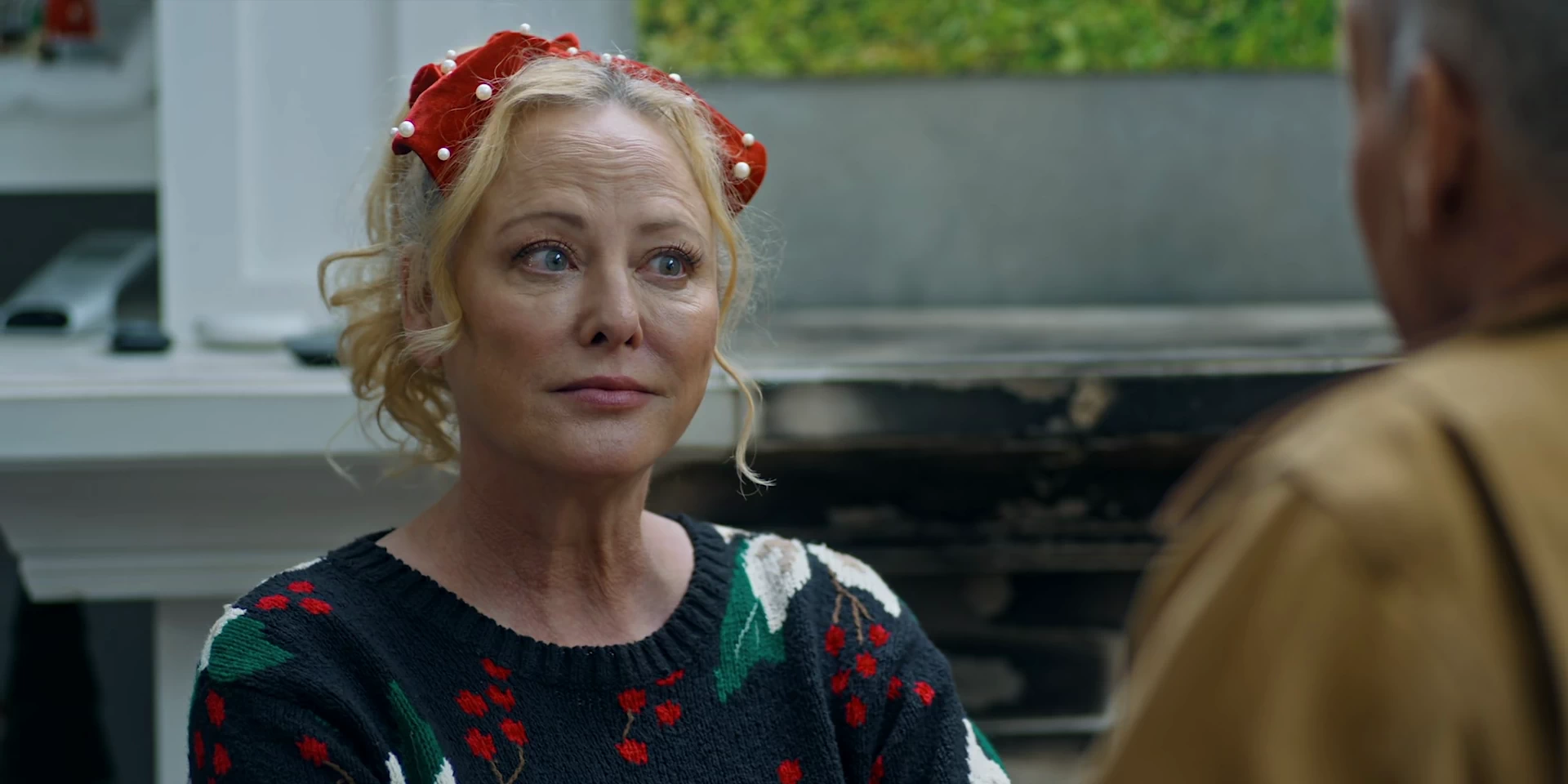 Virginia Madsen as Connie Manetti-Hanahan in Holidazed. She is wearing a festive black sweater adorned with red cherries and green leaves, along with a red headband decorated with pearls. Her blonde hair is loosely styled with soft curls framing her face. She has a thoughtful, slightly amused expression as she listens to someone off-camera. The background shows a modern kitchen with a countertop and some appliances, while a blurred male figure in a brown jacket is partially visible in the foreground