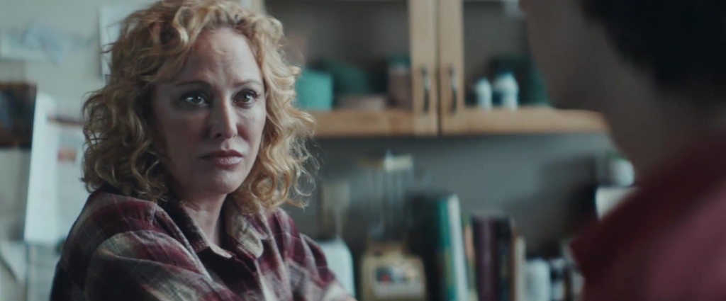 Virginia Madsen as Abigail Charm in All the Wilderness. She stands in a kitchen, wearing a plaid shirt, her curly blonde hair framing her face. Her expression is serious and emotional as she looks at someone in the foreground, it's a tense and heartfelt conversation.