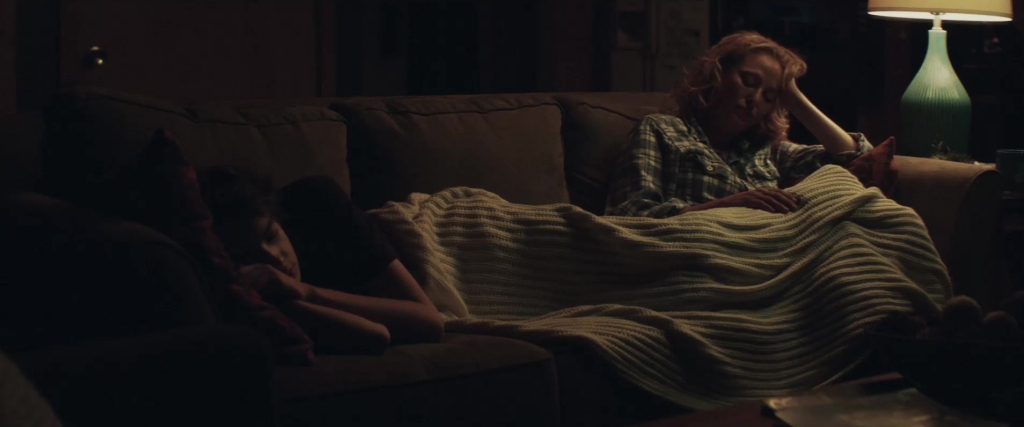 Virginia Madsen as Abigail Charm in All the Wilderness. She is sitting on a dimly lit couch, wrapped in a striped blanket, wearing a plaid shirt. With one hand resting against her head, she appears deep in thought or exhaustion.