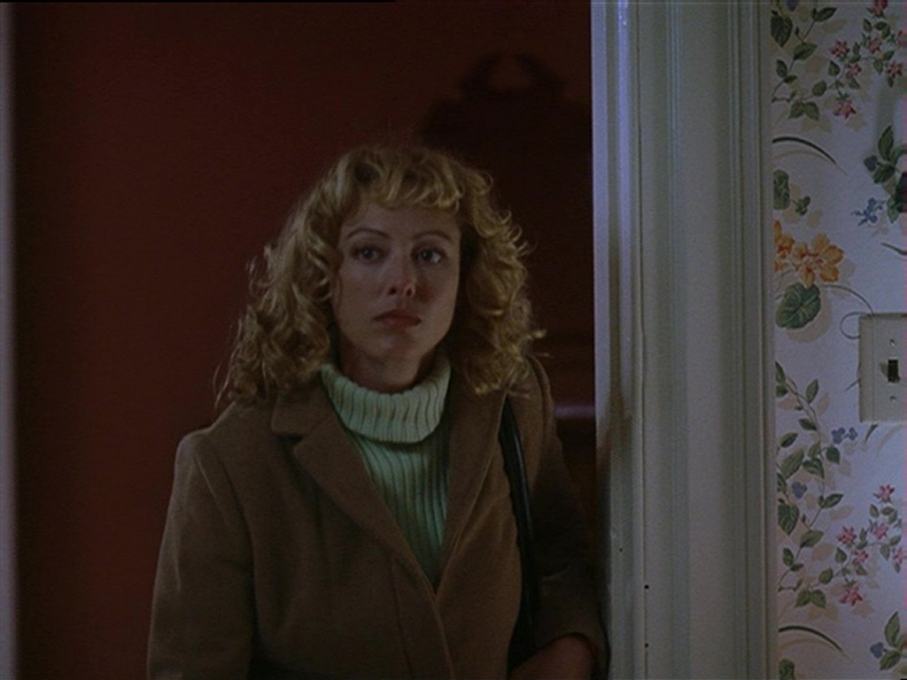 Virginia Madsen as Penny Tillman in American Gun. She stands in a doorway, wearing a camel-colored coat over a green turtleneck sweater, her curly blonde hair framing her face. Her expression is serious and uncertain as she looks into the dimly lit room. The contrast between the warm red interior and the floral wallpaper in the foreground adds a sense of tension to the scene.
