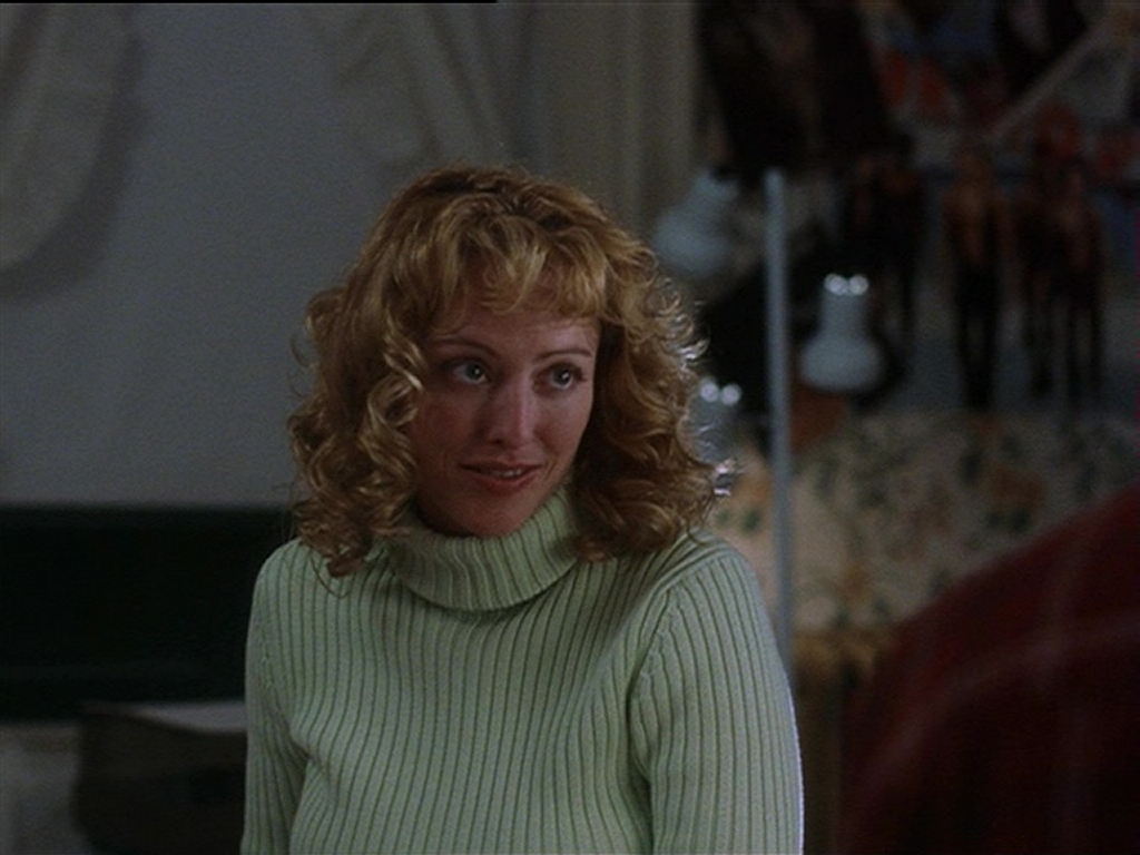 Virginia Madsen as Penny Tillman in American Gun. She wears a light green turtleneck sweater, her curly blonde hair framing her face. She has a slightly amused or skeptical expression as she looks at someone off-camera. The warmly lit room in the background, with various objects and decorations, suggests a cozy yet lived-in space