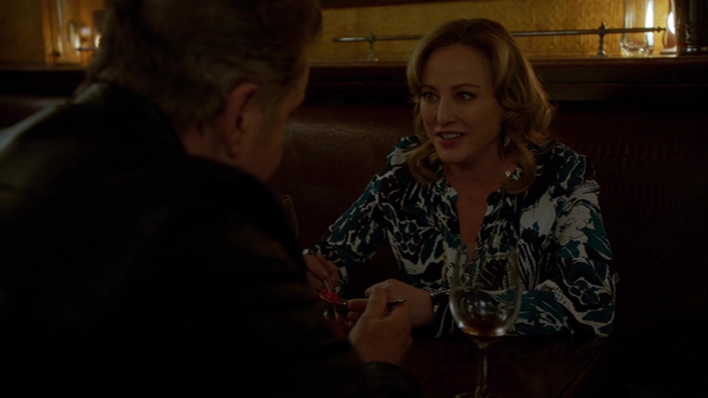Virginia Madsen as Paige Cowan in Elementary, sitting at a dimly lit bar or restaurant, engaged in an intense conversation with a man across from her. She wears a stylish patterned blouse and has a confident yet serious expression. A glass of wine sits in front of her, adding to the intimate and tense atmosphere of the scene.