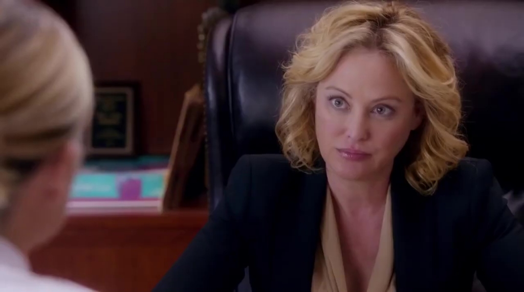 Virginia Madsen as Judy Hammer in Hornet's Nest. She sits behind a desk in a black blazer and beige blouse, giving a serious yet composed expression. The office setting suggests a professional or investigative atmosphere.
