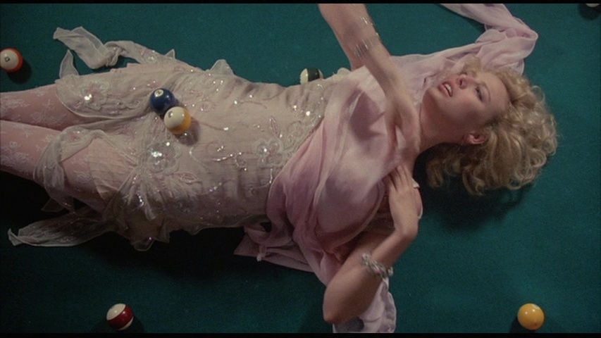 Virginia Madsen lies dramatically on a green pool table, surrounded by scattered billiard balls. She wears a sparkling, sheer pink dress with lace details and flowing fabric, evoking an ethereal, dreamlike aesthetic. Her voluminous blonde curls frame her face as she gazes upwards, her expression caught between dazed and glamorous, embodying the neon-soaked decadence of the '80s.