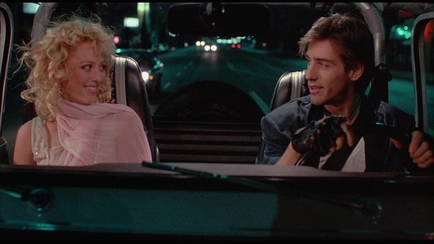 A nighttime scene from Modern Girls (1986). Virginia Madsen, with voluminous curly blonde hair, wears a light pink, semi-sheer outfit with a flowing scarf, sitting in the passenger seat of a Jeep. She smiles playfully at the driver, a young man with tousled brown hair, dressed in a dark jacket, fingerless gloves, and a bandana around his neck. The neon-lit city streets blur in the background, evoking an '80s urban nightlife aesthetic.