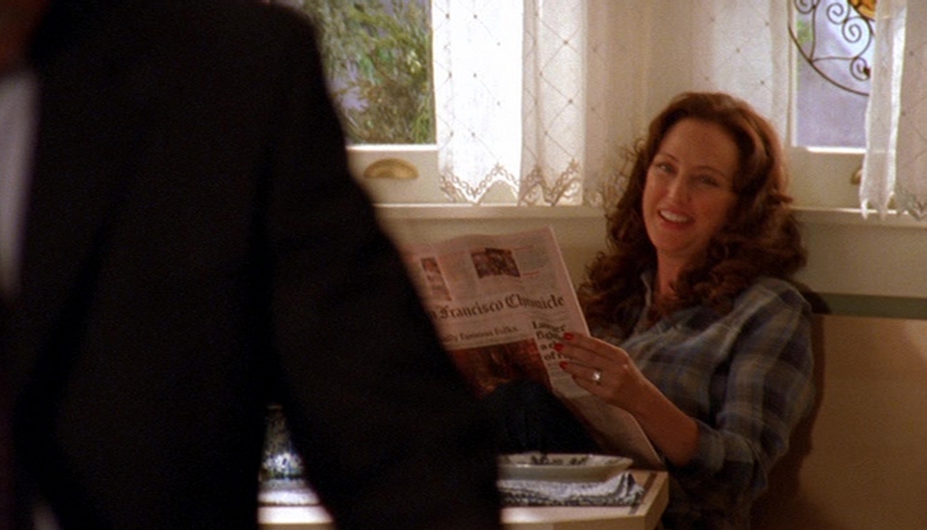 Virginia Madsen as T.J. Jensen in Monk, sitting at a cozy kitchen nook with a smile while reading the San Francisco Chronicle. She wears a plaid shirt, her wavy brown hair framing her face.