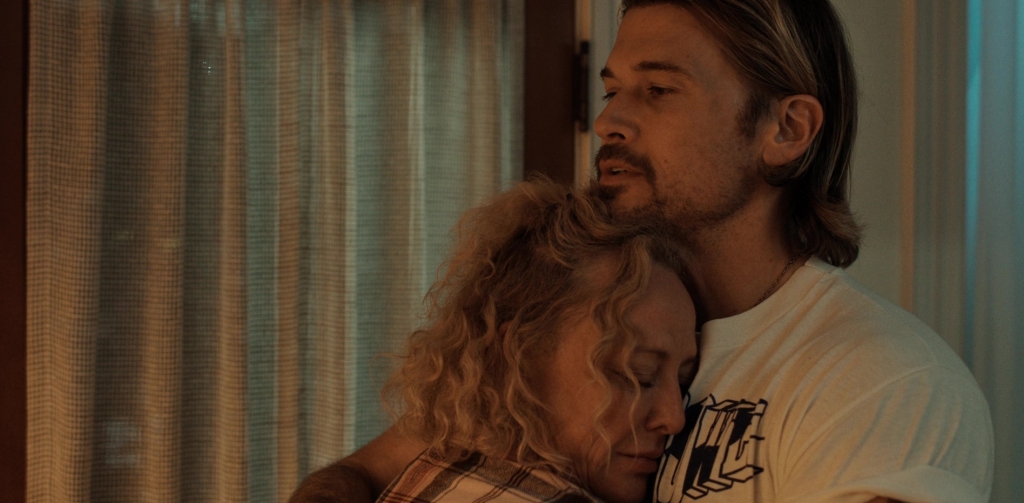 Virginia Madsen as Marge McKnight in Obliterated, resting her head against a man’s chest in an emotional embrace. She wears a plaid shirt, her curly blonde hair slightly disheveled. The man, wearing a white graphic t-shirt, has a solemn expression as he holds her close. The dimly lit room features a softly illuminated curtain in the background, adding to the intimate and comforting atmosphere