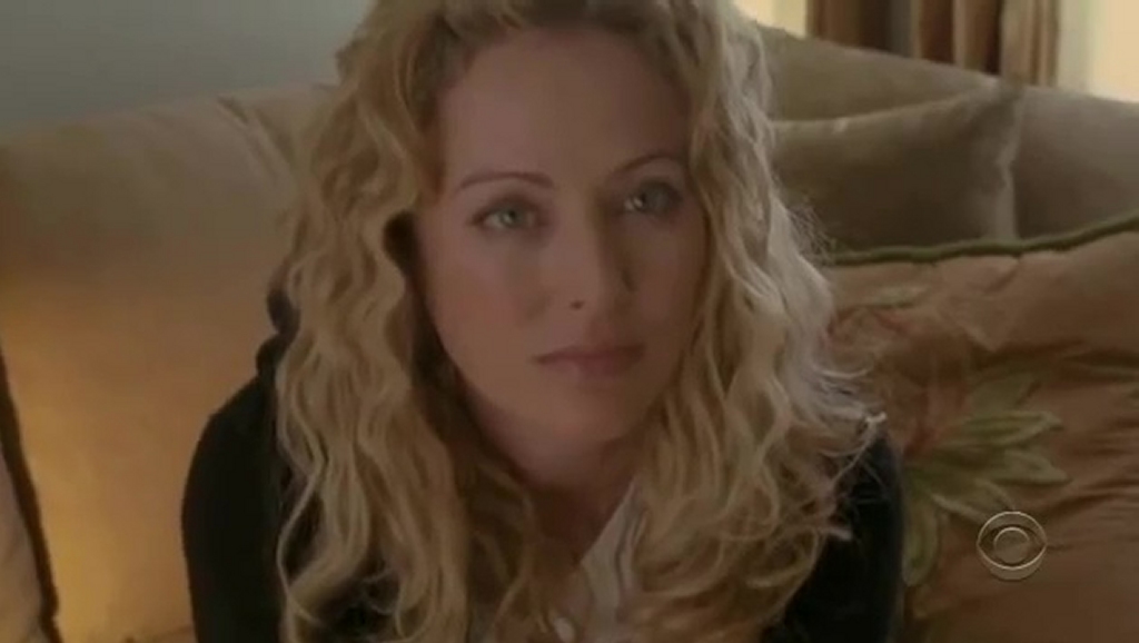 Virginia Madsen as Hope Stevens in Smith, sitting on a couch with an intense gaze. Her long, curly blonde hair frames her face, and she wears a black jacket over a white top. The dim lighting and warm-toned pillows in the background add to the moody atmosphere.