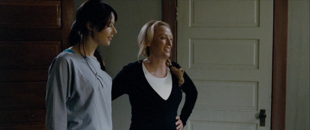 Virginia Madsen in The Haunting in Connecticut, wearing a black cardigan over a white top, standing beside another woman. She smiles warmly with her hand on her hip in a dimly lit room with wooden doors.