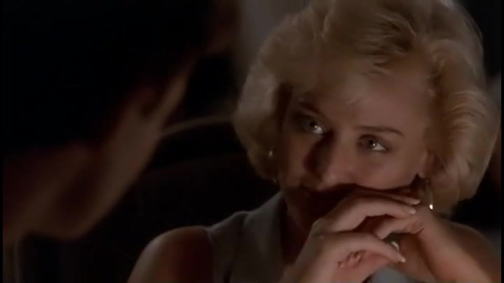 Virginia Madsen as Anne Scholes in Third Degree Burn (1989), gazing thoughtfully at someone off-screen. Her blonde hair is elegantly styled, and she rests her chin on her hands, her expression both alluring and mysterious. The dim lighting enhances the film’s moody, noir-like atmosphere.