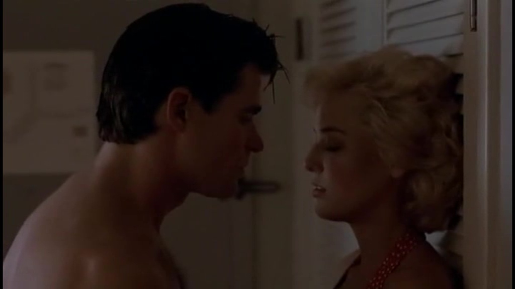 Virginia Madsen as Anne Scholes in Third Degree Burn (1989), leaning against a wall in a dimly lit room. She wears a red halter top, her blonde hair tousled, as she faces a shirtless Treat Williams. Their close proximity and intense expressions add to the film’s sultry, suspenseful tone.
